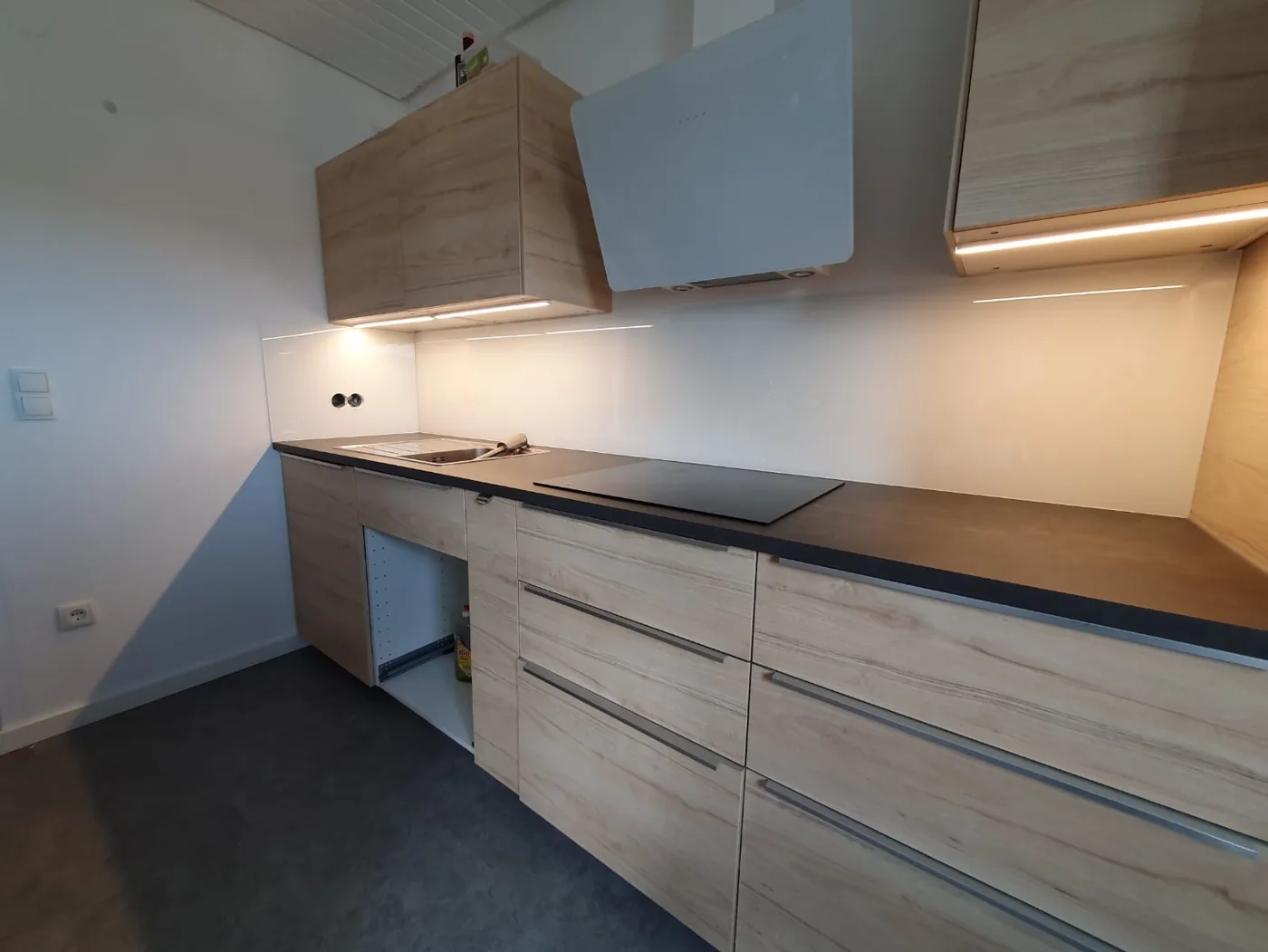 Weiße Glasrückwand in einer Küche mit Holzdekor, mit Ausschnitten für Steckdosen, unter beleuchteten Hängeschränken